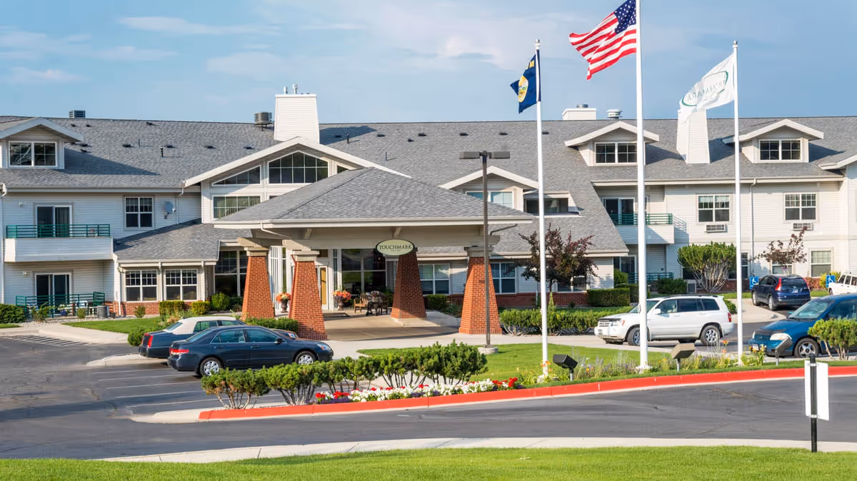 Exterior view of Touchmark on Saddle Drive, a multi-story senior living facility with a covered entrance supported by brick pillars. Several cars are parked in front, and three flagpoles display the American flag, a state flag, and a facility flag. The building has multiple windows, balconies, and a gray shingled roof under a partly cloudy sky.