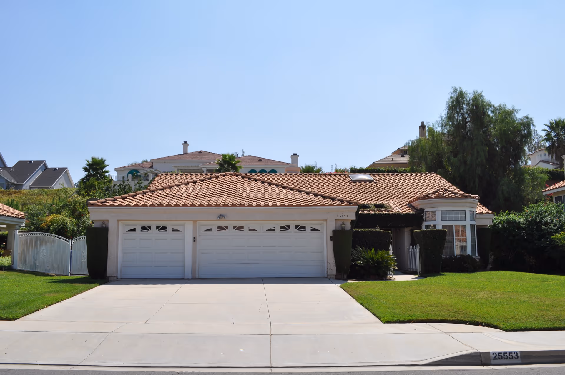 Front exterior view of a single-story house with a red tiled roof, three white garage doors, neatly trimmed bushes, green lawn, and a clear blue sky.
