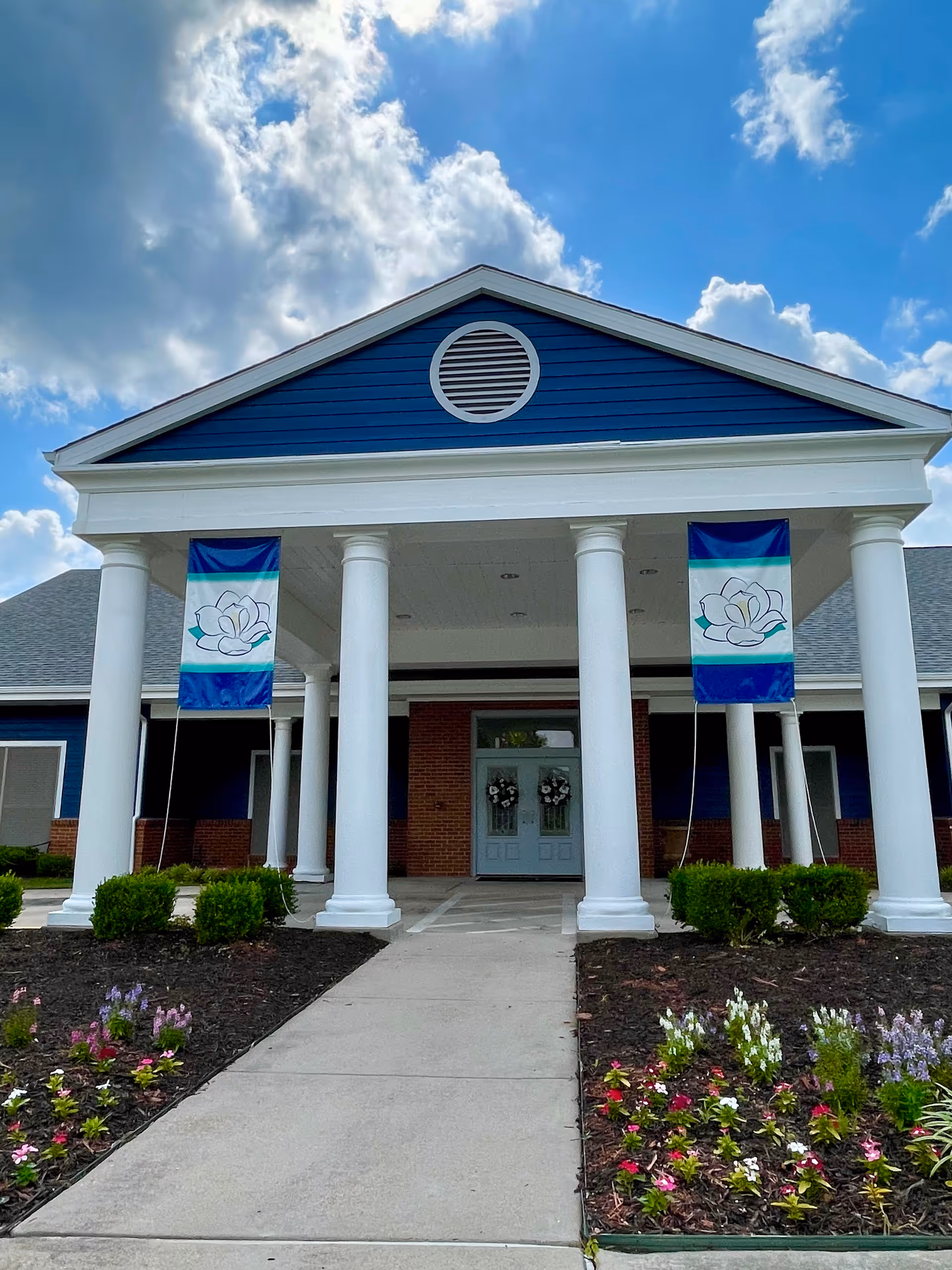 Front exterior view of Magnolia Assisted Living facility showing a blue building with white columns, two flags with a lotus flower design, a concrete walkway, and flower beds on either side under a partly cloudy sky.