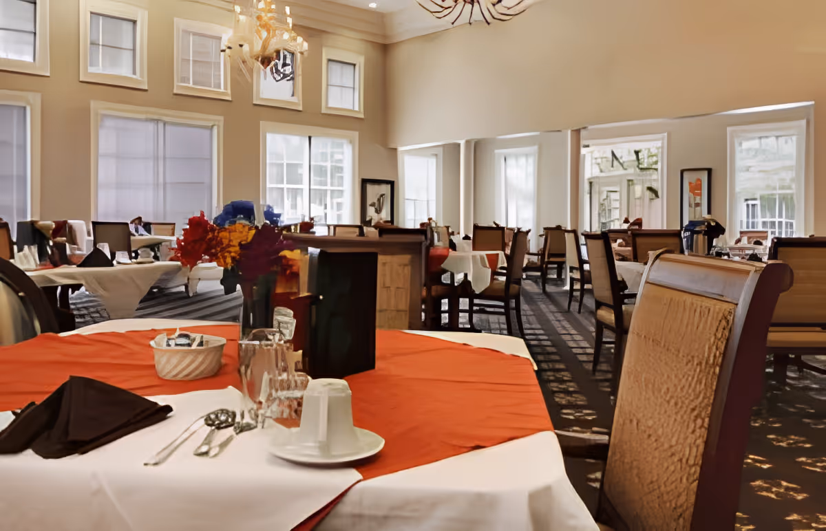 Bright dining room with tables set for meals, chairs, high ceilings and large windows.