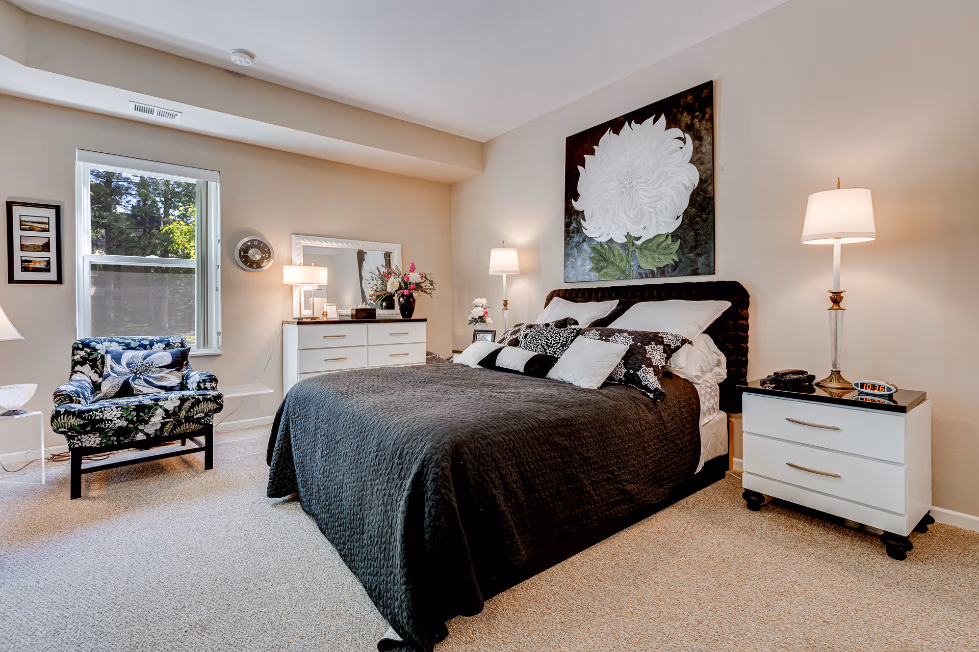 A cozy bedroom with a large bed covered in a dark quilt and multiple pillows. There is a floral armchair near a window with a view of trees outside. The room features two white dressers with lamps and decorative items, including a large floral painting above the bed. The walls are light-colored and the floor is carpeted.