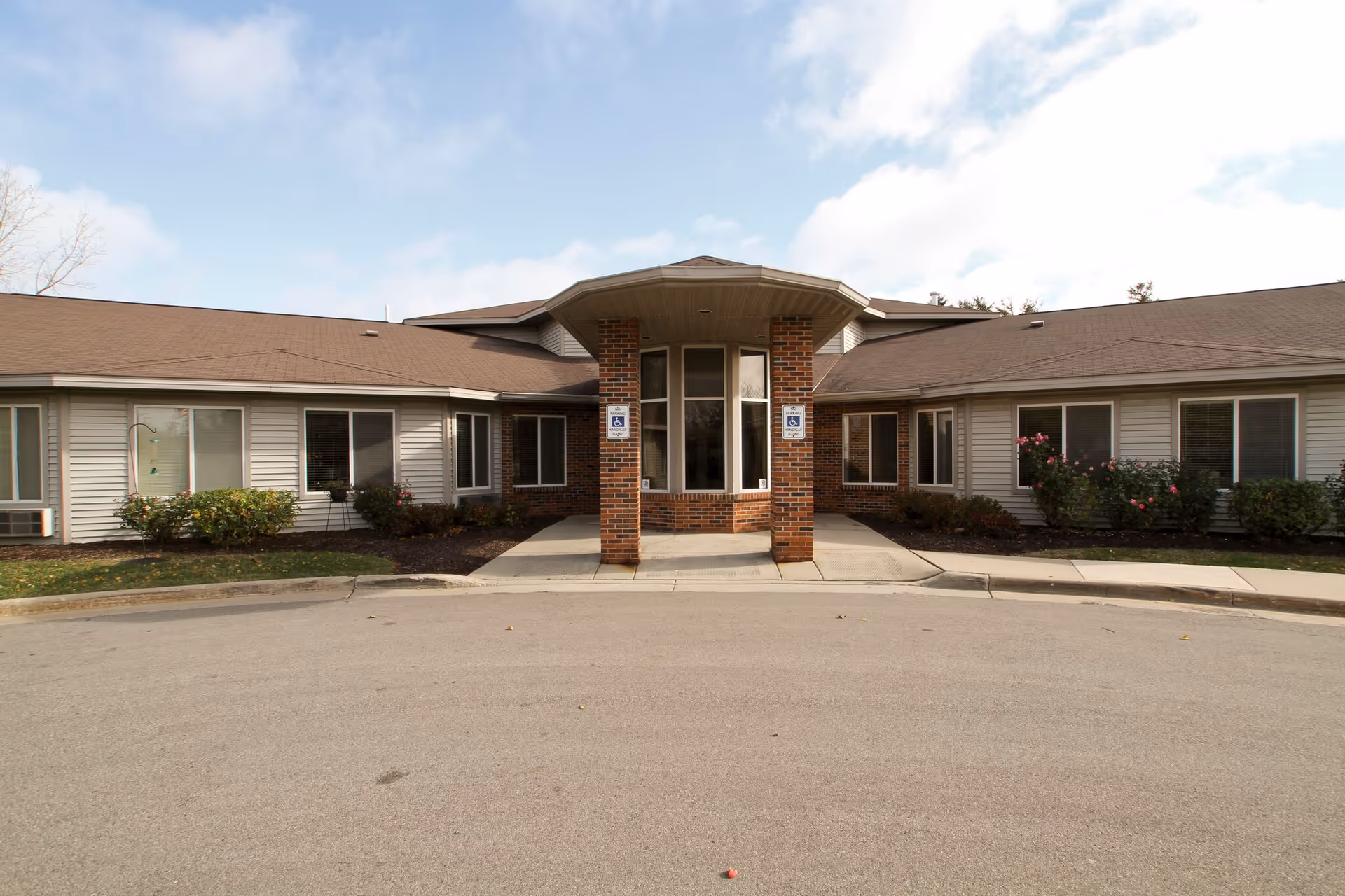 Front exterior view of a single-story building with a covered entrance supported by two brick pillars. The building has a brown shingled roof, beige siding, and several windows with blinds. There are small bushes and flowers planted along the front of the building. Two handicap parking signs are visible near the entrance.