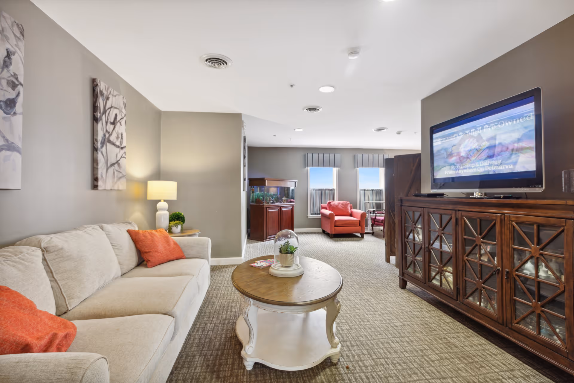 A cozy living room in an assisted living facility featuring a beige sofa with orange pillows, a round wooden coffee table with a small plant and magazines, a wooden cabinet with a flat-screen TV on top, and a red armchair near two windows with blue valances. The room has neutral-colored walls and carpeted flooring.