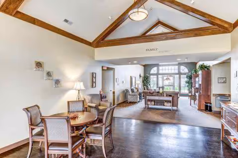 A spacious and well-lit common area in a senior living facility featuring a round wooden table with four chairs in the foreground, comfortable armchairs, a wooden coffee table, and large windows letting in natural light. The ceiling has exposed wooden beams and a modern light fixture. The room is decorated with plants, wall art, and a sign that reads 'FAMILY' above the entrance to another seating area.