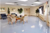 A spacious, well-lit common area with several beige armchairs arranged around a wooden coffee table. The room has light blue flooring, white walls, and large windows with curtains allowing natural light to enter. There are framed pictures and potted plants decorating the space.