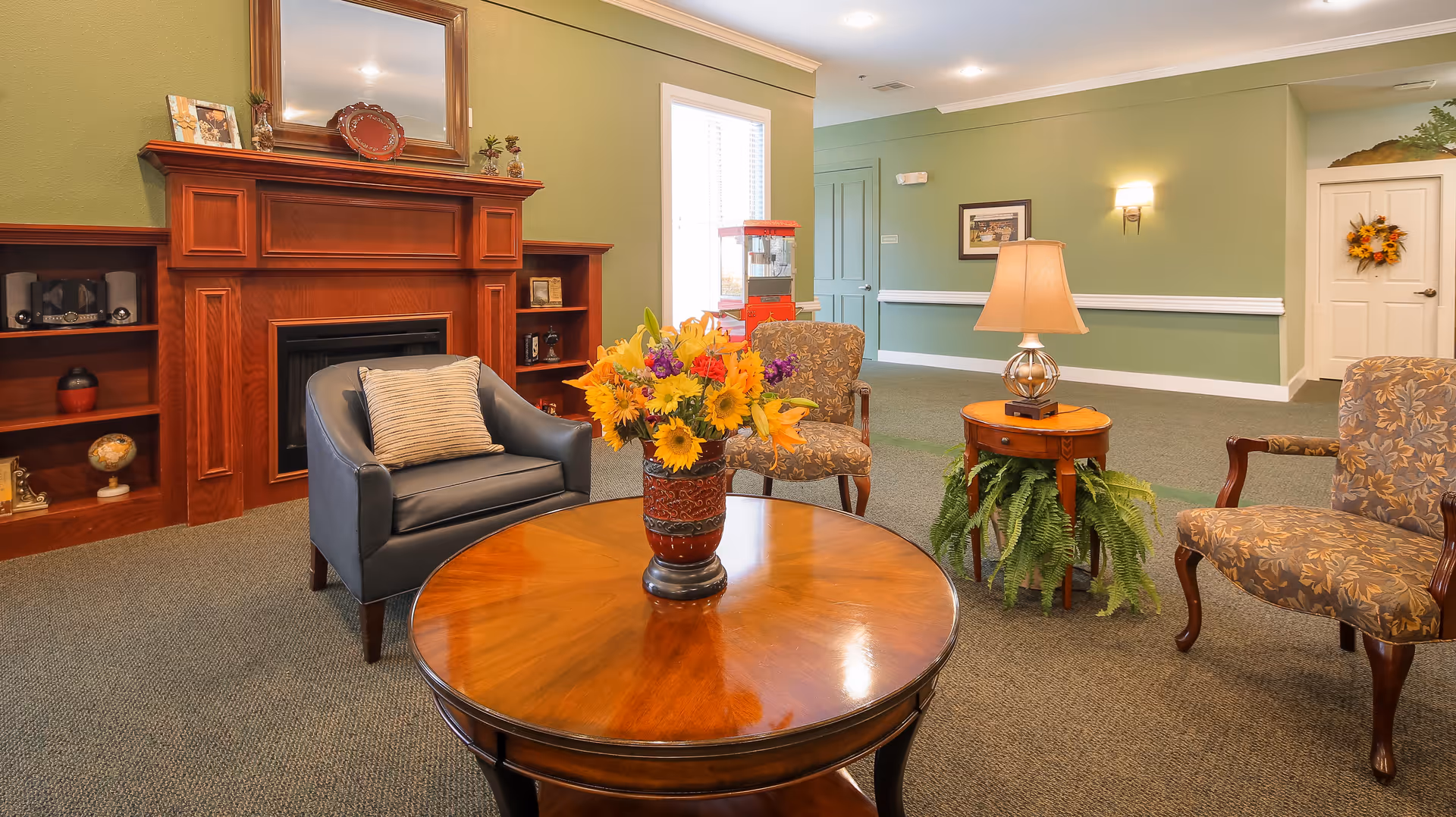 A cozy living room area with a wooden round table in the center holding a vase of colorful flowers. Surrounding the table are three chairs: one dark leather armchair with a pillow and two patterned fabric armchairs. In the background, there is a wooden fireplace mantel with decorative items and a mirror above it. The walls are painted green with white trim, and there is a side table with a lamp and a potted fern underneath. A door and framed picture are visible on the far wall.