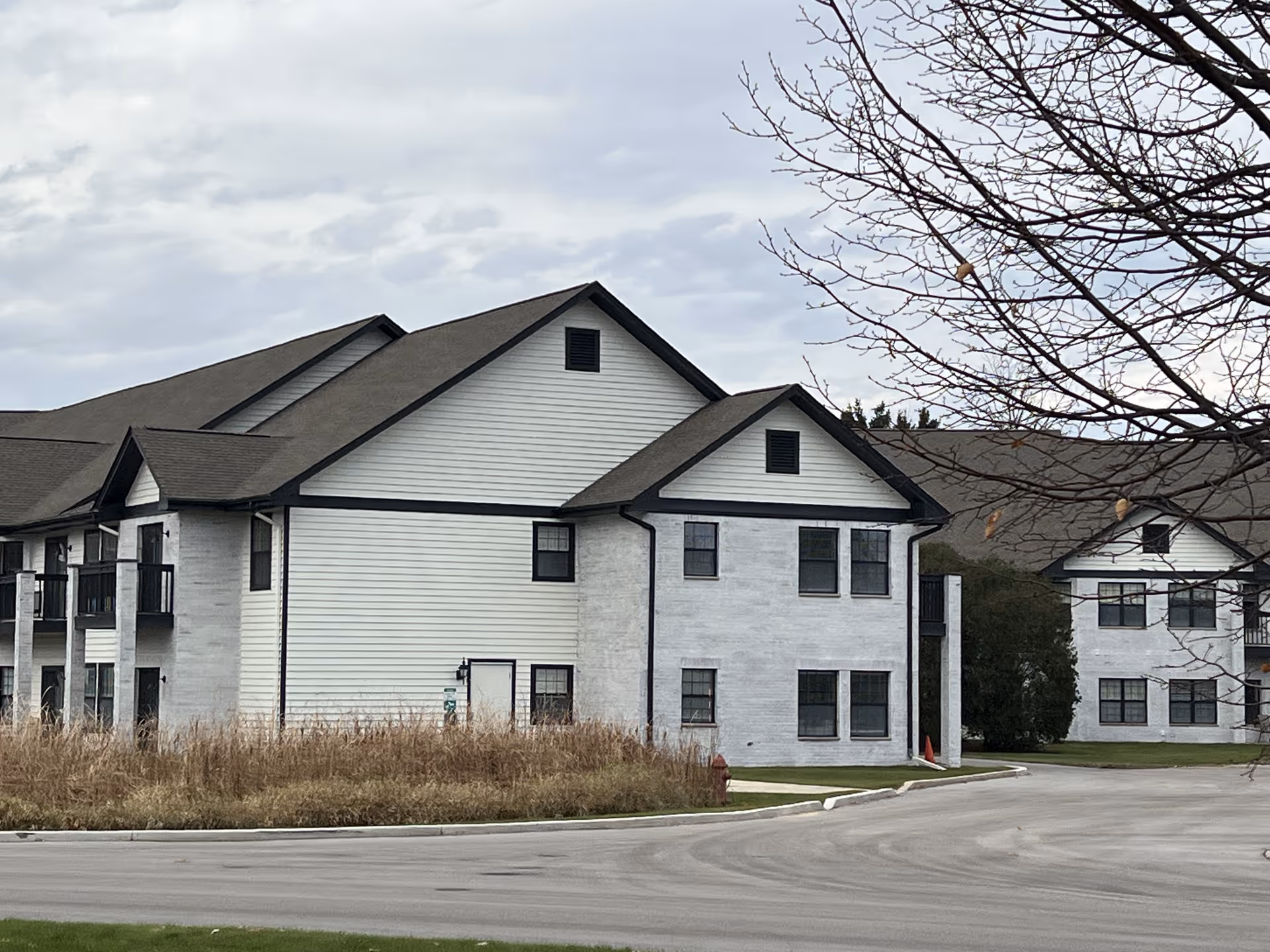 Exterior view of a two-story senior living facility building with white siding and black trim, surrounded by a paved driveway and some leafless trees under a cloudy sky.
