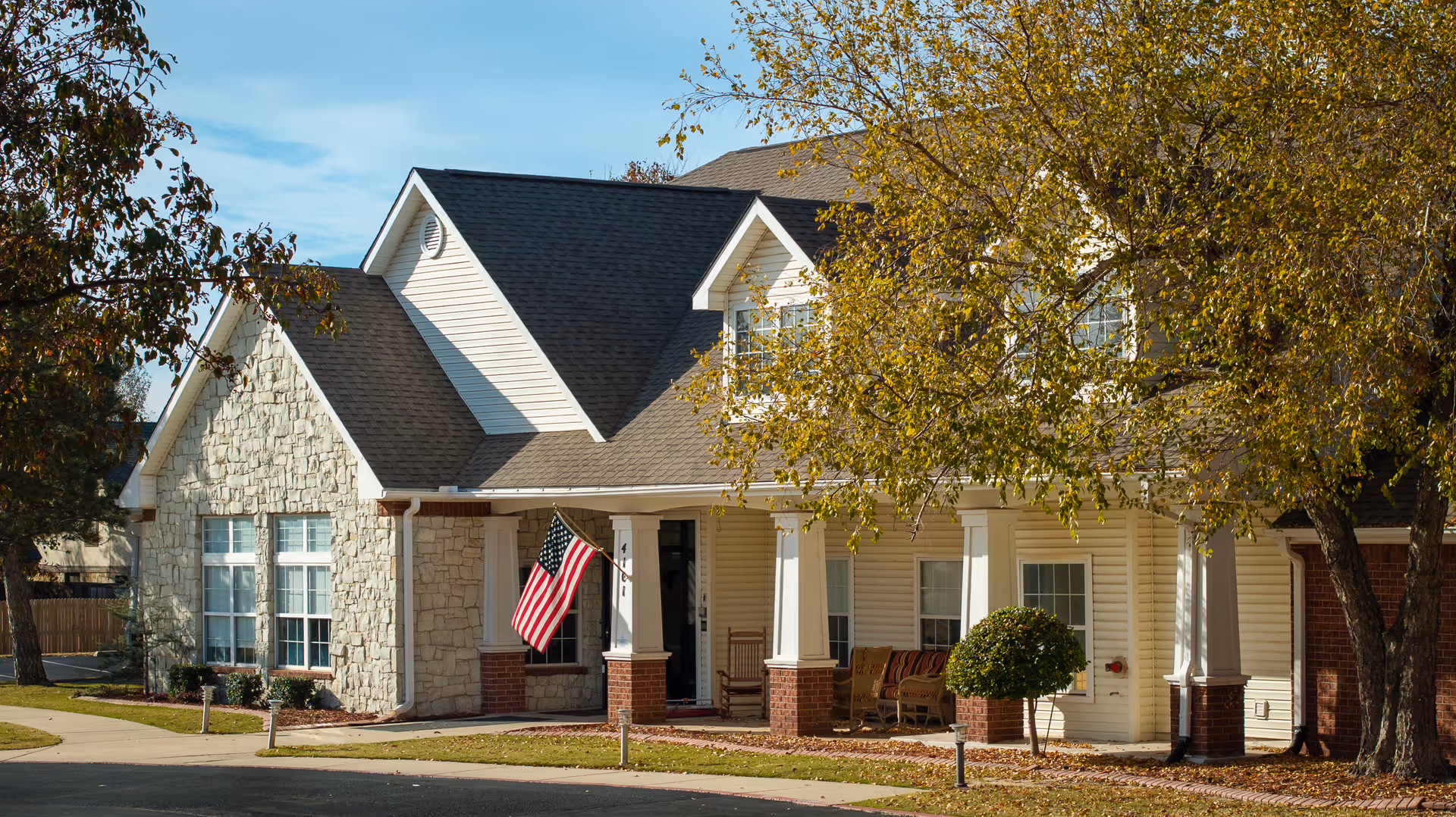 Exterior view of a residential-style assisted living facility with beige siding, stone accents, a covered porch with rocking chairs, an American flag hanging near the entrance, and trees with autumn foliage.