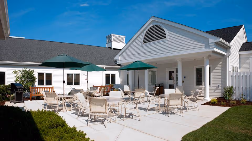 Outdoor patio area at Edgewood LifeCare Community with multiple tables and chairs arranged on a concrete surface. Green umbrellas provide shade over some tables. The building exterior is white with a dark roof, and there is a grill and some benches near the windows. The sky is clear and blue.