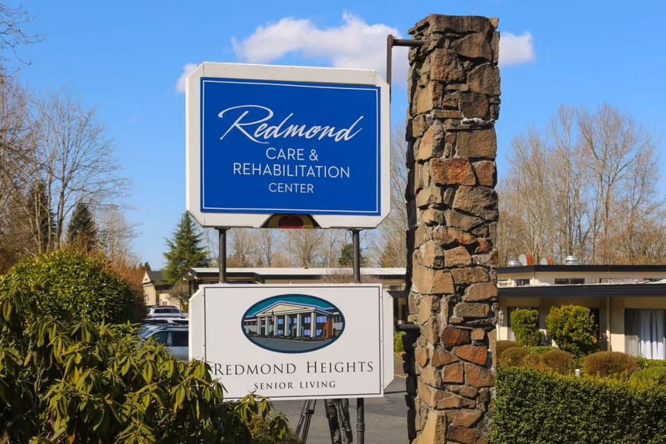 Outdoor view of the entrance area of Redmond Care & Rehabilitation Center and Redmond Heights Senior Living, featuring two signs mounted on metal poles next to a tall stone pillar, with bushes and trees in the background under a clear blue sky.