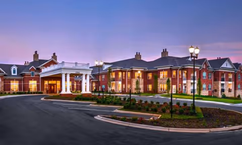 Front exterior of Waltonwood Providence, brick building with circular driveway and lit lampposts at dusk.