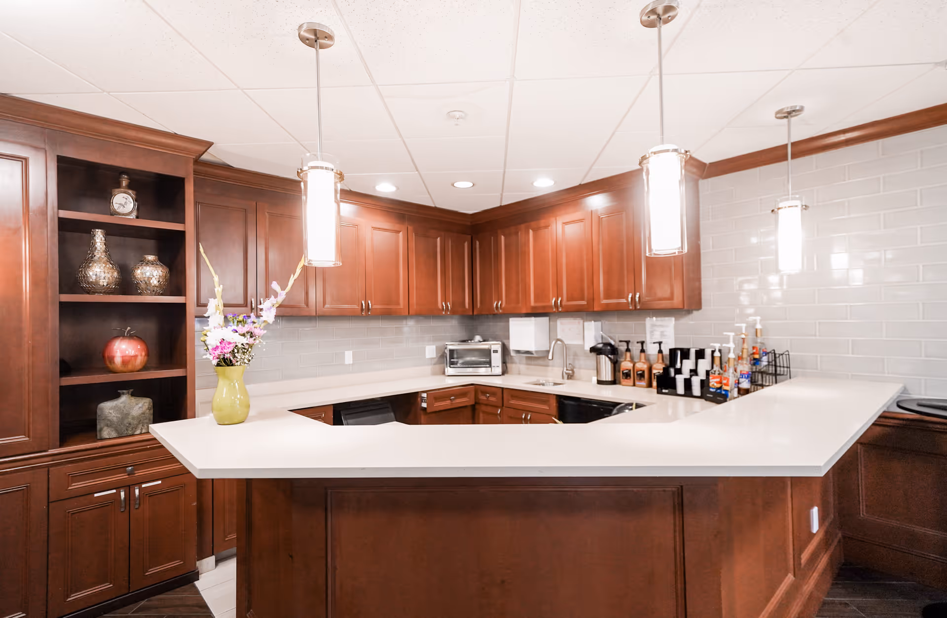 A modern kitchen area with dark wooden cabinets, white countertops, and a white tiled backsplash. The kitchen features a corner sink, a toaster oven, and various coffee and syrup dispensers on the counter. Three pendant lights hang from the ceiling, illuminating the space. A vase with flowers is placed on the counter near open shelving with decorative items.