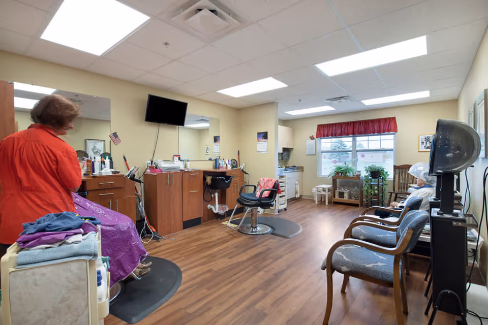 Interior of a hair salon room in a senior living facility with wooden flooring, beige walls, and fluorescent ceiling lights. There are salon chairs, a hair dryer, a counter with hair products, and a TV mounted on the wall. A woman in a red jacket is standing near a salon chair covered with a purple cape, and an elderly person is seated under a hair dryer near the window with red curtains and plants on a small table.