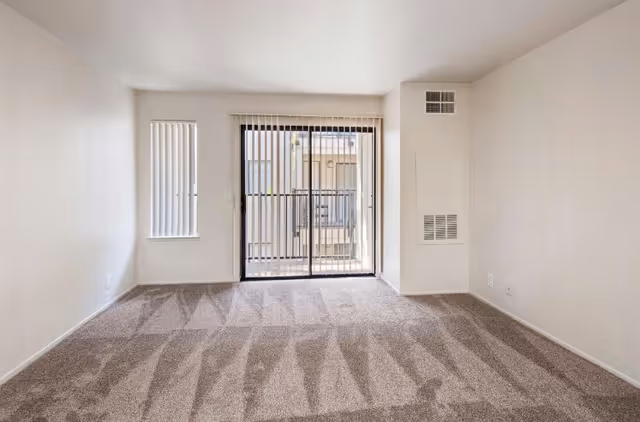 Empty room with beige carpet and white walls featuring a small window with vertical blinds on the left and a sliding glass door with vertical blinds leading to a balcony or outdoor area.
