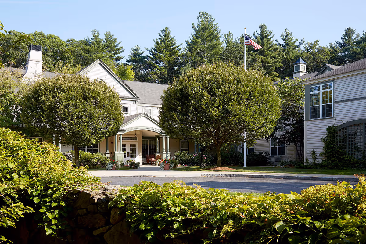 Entrance of a residential building with a covered porch, manicured trees and landscaped greenery under a clear sky.