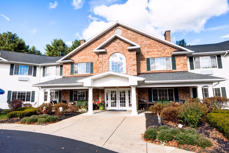 Front exterior view of a two-story senior living facility with a brick facade in the center and white siding on the wings. The entrance has a covered porch supported by white columns, with glass double doors. There are shrubs and landscaped plants along the walkway leading to the entrance, and a clear blue sky with some clouds above.