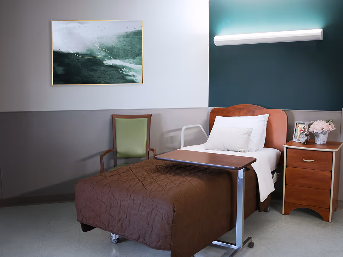 A single hospital-style bed with a brown quilted blanket and white pillows in a room. Next to the bed is a wooden bedside table with three drawers, a framed photo, an alarm clock, and a vase of pink flowers. A green cushioned chair is positioned against the wall. Above the chair hangs a framed abstract painting with green and white tones. The walls are painted in two shades, with a darker green section behind the bed and a lighter gray on the other walls. A horizontal light fixture is mounted above the bed.