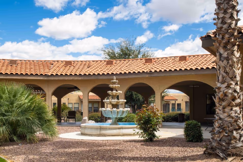 Outdoor courtyard area with a multi-tiered stone fountain in the center, surrounded by desert landscaping including palm trees and bushes. The courtyard is bordered by a building with a red tile roof and arched walkways under a partly cloudy blue sky.