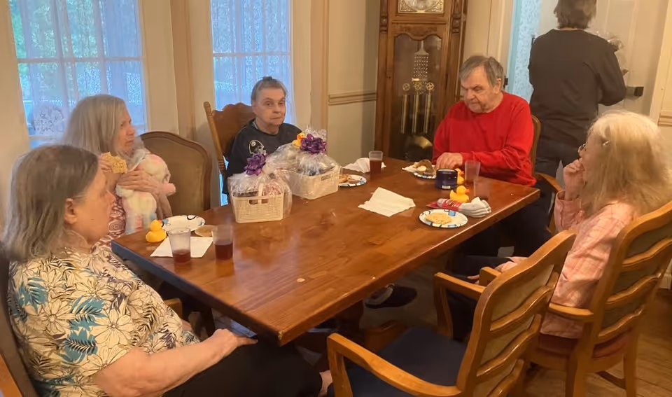 Five elderly individuals sitting around a wooden dining table in a room with large windows covered by sheer curtains. The table has plates with snacks, cups with drinks, and two gift baskets wrapped with purple ribbons. One person is standing near the wall in the background.
