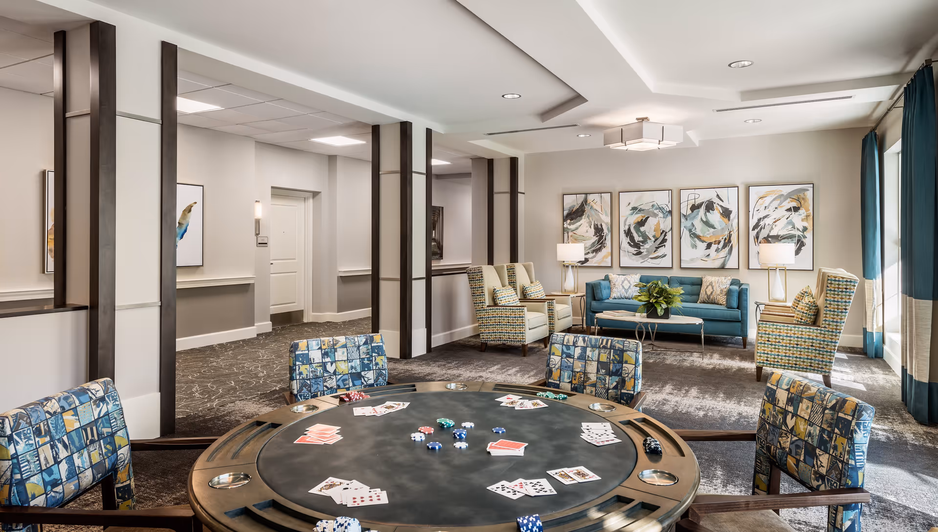 A well-lit common area in an assisted living facility featuring a round poker table with playing cards and poker chips, surrounded by patterned chairs. In the background, there is a seating area with a teal sofa, two patterned armchairs, side tables with lamps, and abstract artwork on the wall. Large windows with teal curtains allow natural light to fill the room.