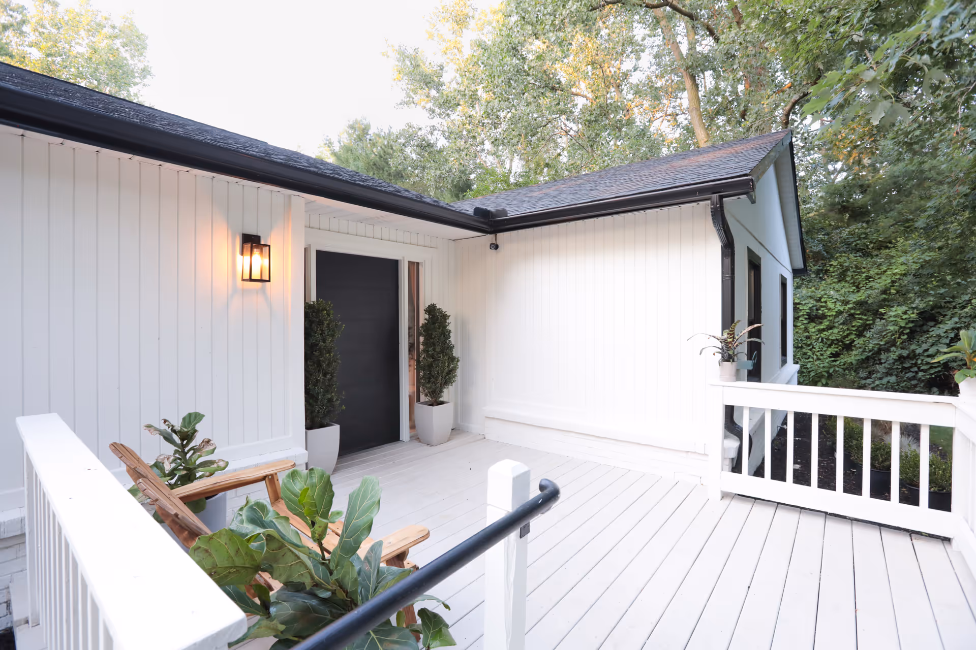 A white wooden deck area outside a building with a black door, two potted plants flanking the door, a wall-mounted light fixture, wooden chairs, and surrounding greenery.