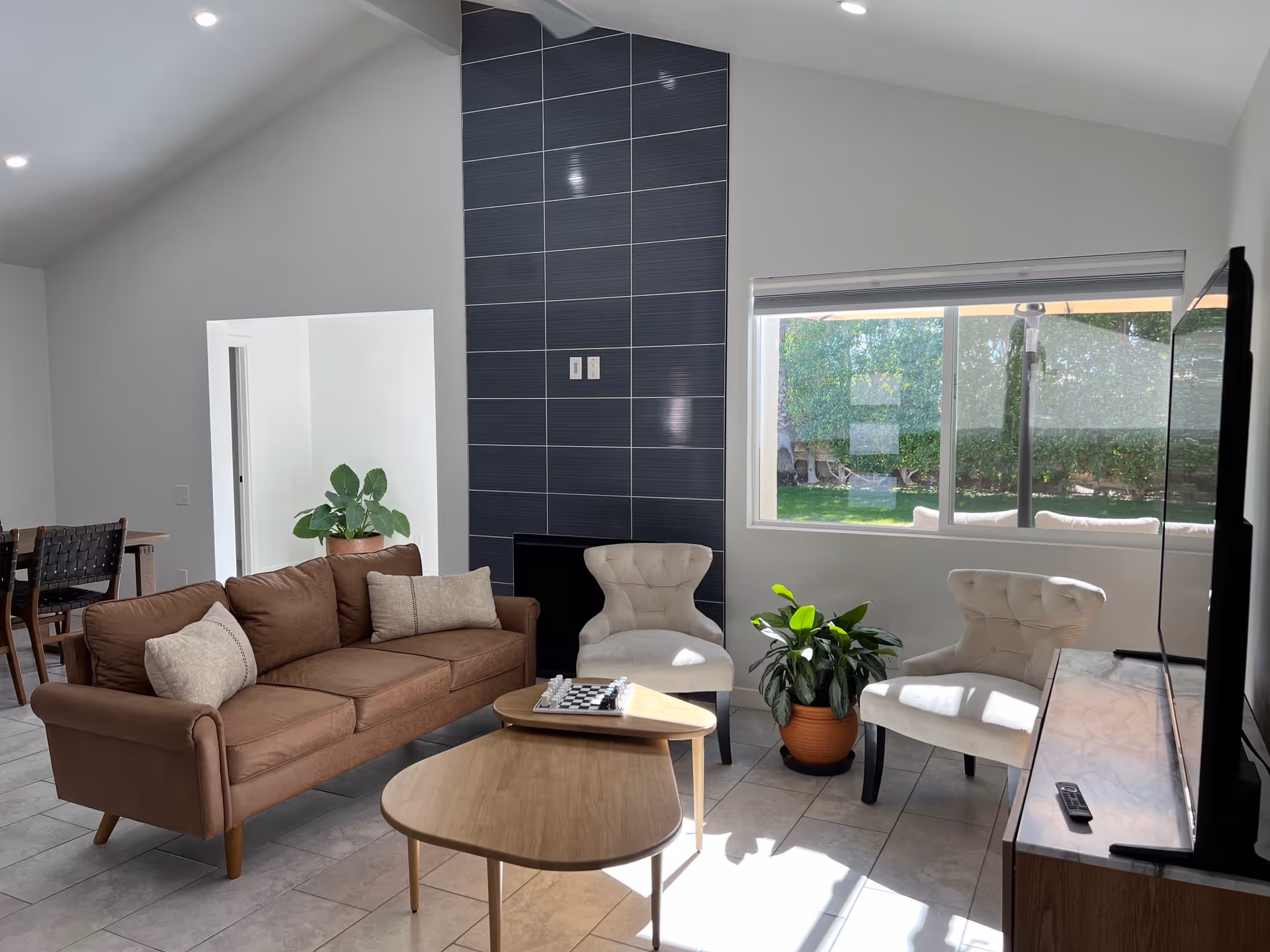 Bright modern living room with a brown sofa, two beige armchairs, coffee tables, a potted plant, and a large window looking out to a yard.