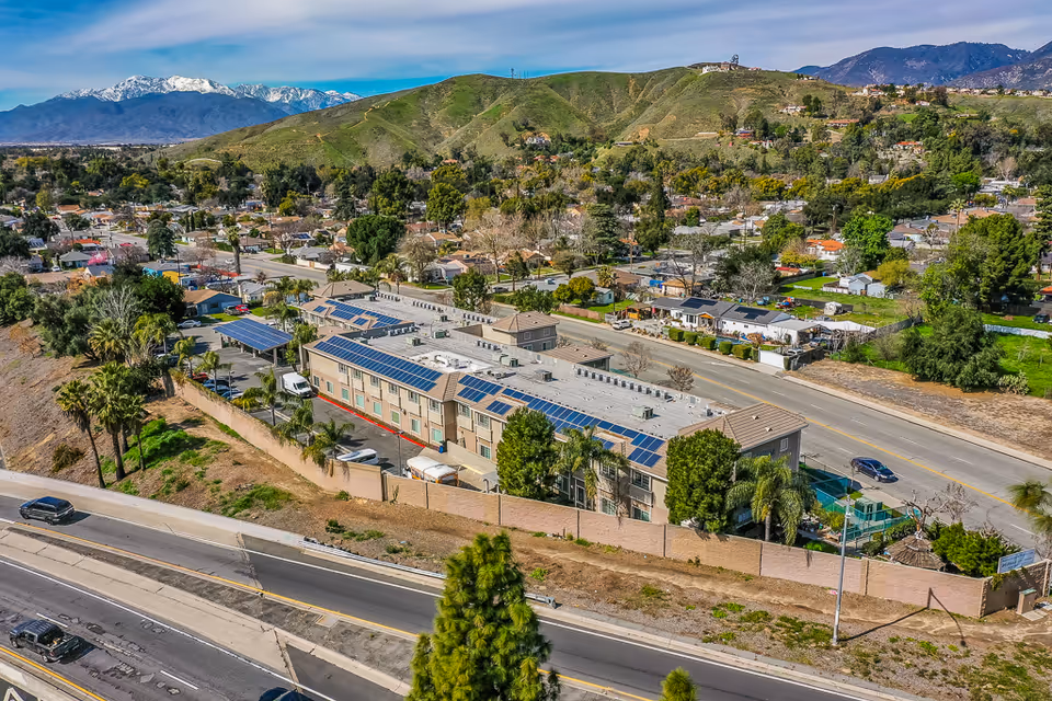 Aerial view of a multi-story senior living building with solar panels on the roof, surrounded by roads, trees, and a residential neighborhood with hills in the background.