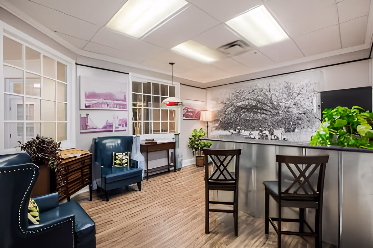 A cozy senior living common area with wooden flooring, two dark blue armchairs with decorative pillows, a small wooden table, and a counter with two wooden chairs. The walls feature framed artwork and a large black and white mural of a tree. There are also green plants adding a touch of nature to the space.