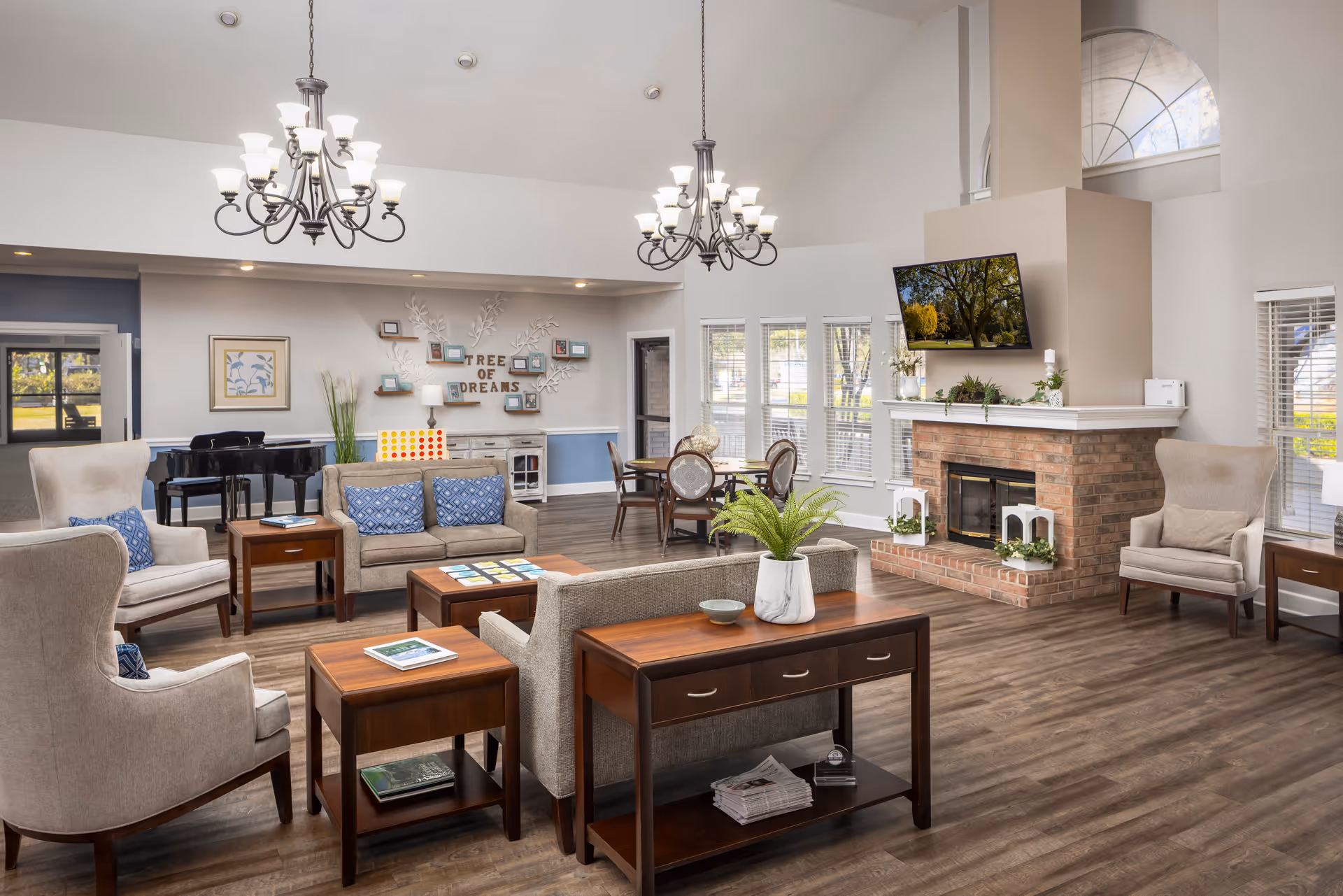 A spacious and well-lit living room area in a senior living facility featuring comfortable armchairs and sofas with blue cushions, wooden side tables, a brick fireplace with a mounted TV above it, large windows with white blinds, two elegant chandeliers, a piano in the background, and a decorative wall with the words 'Tree of Dreams'.