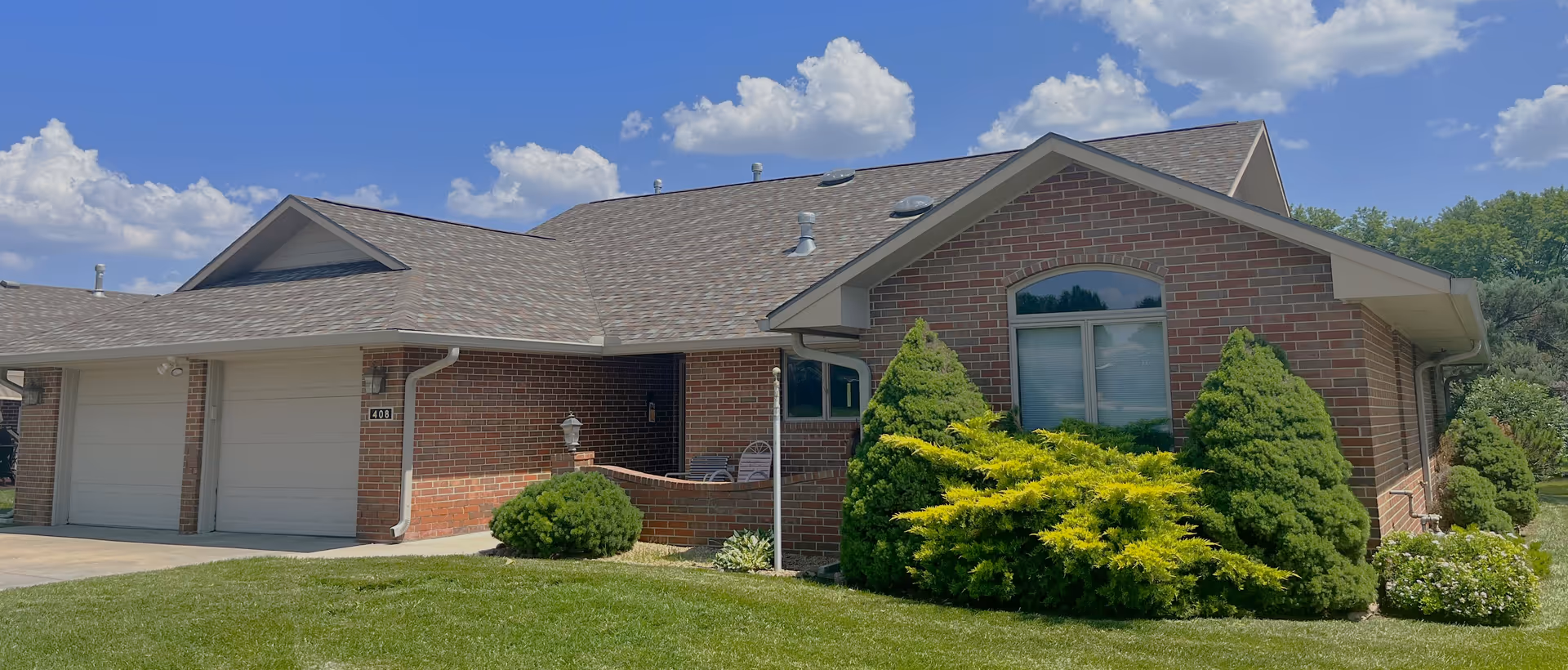 Single-story brick building with an attached two-car garage, manicured shrubs and a lawn under a blue sky.