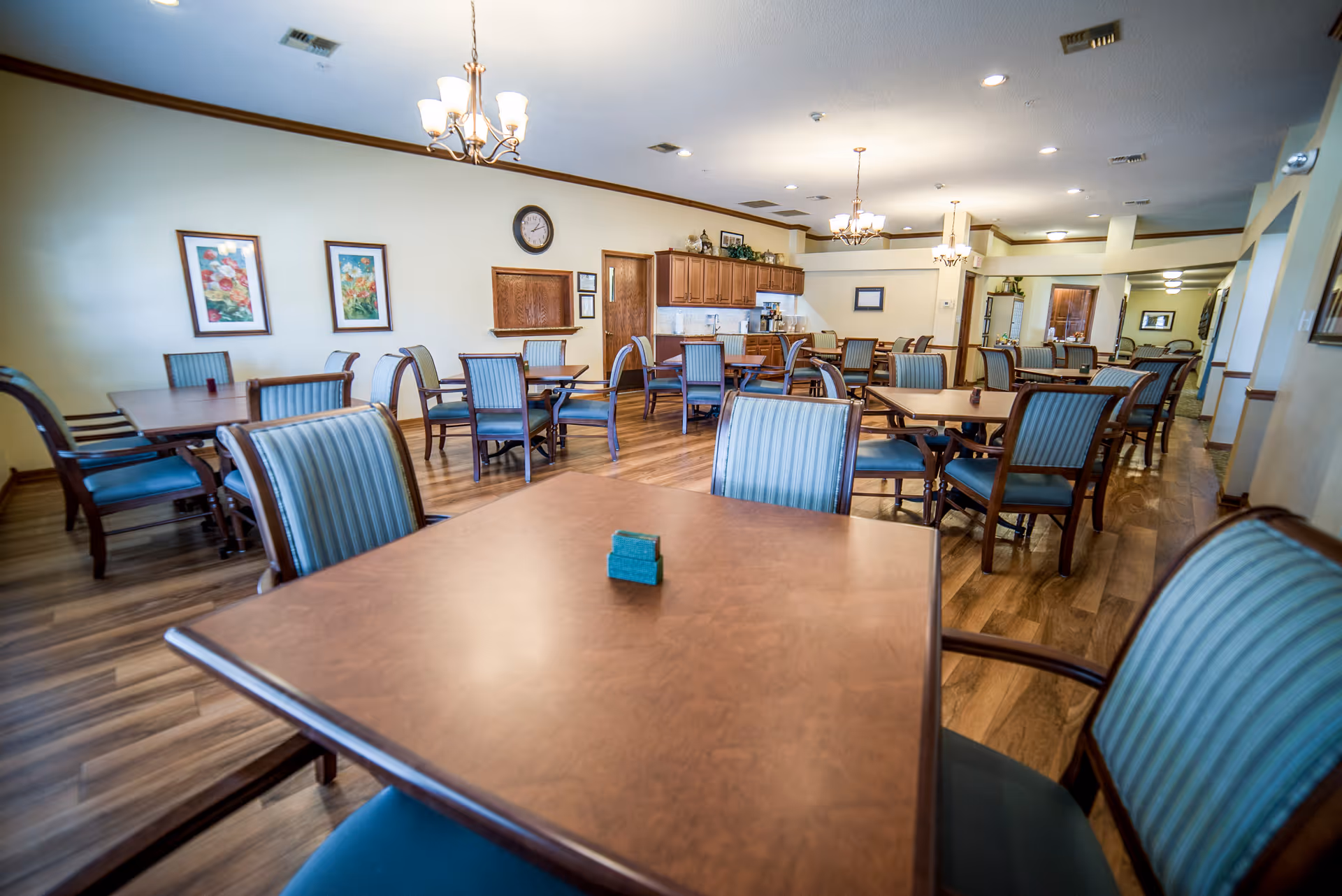 A spacious dining room with multiple wooden tables and chairs with blue striped upholstery. The room has wooden flooring, light-colored walls, framed floral artwork, and ceiling lights including chandeliers. There is a clock on the wall and a counter area with cabinets and a coffee machine in the background.