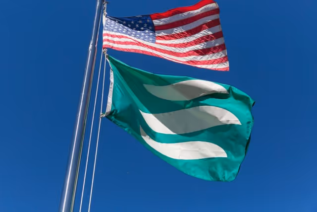 An American flag and a green flag with white wave-like shapes flying on flagpoles against a clear blue sky.