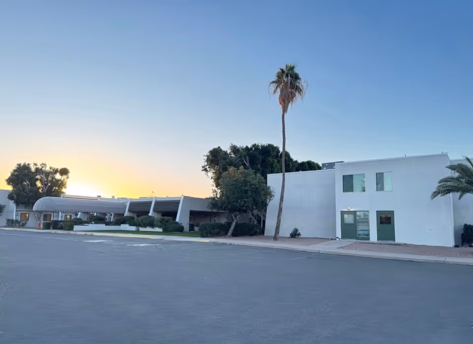 Exterior view of Rio Vista Post Acute And Rehabilitation facility at sunset, showing a white two-story building with green doors and windows, a covered entrance, palm trees, and a large paved parking area in front.
