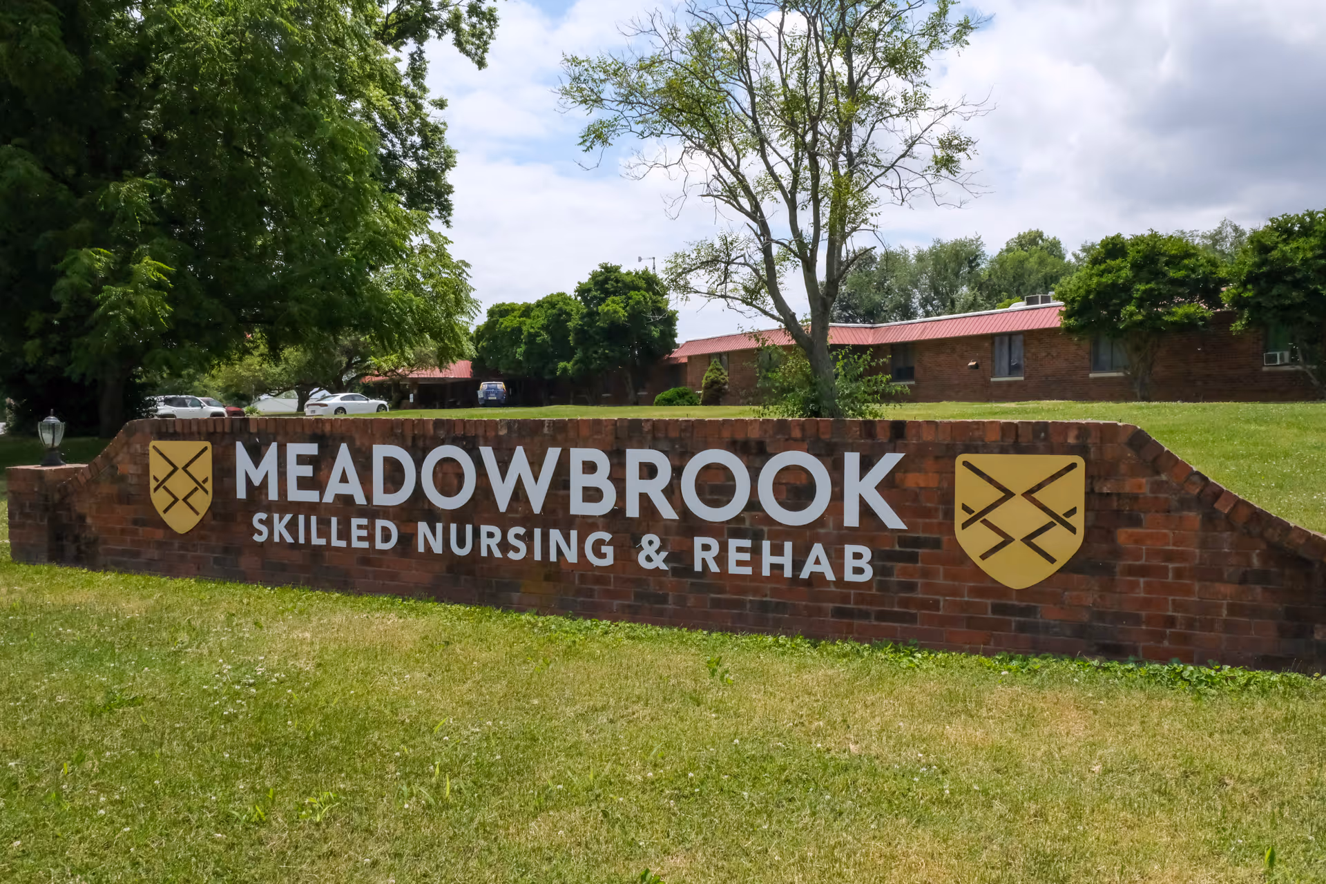 A brick sign for Meadowbrook Skilled Nursing & Rehab situated on a grassy area with trees and a building with a red roof in the background under a partly cloudy sky.