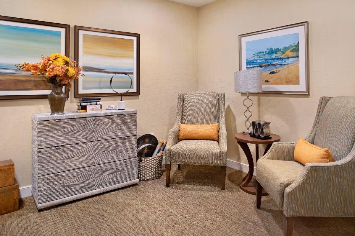 A cozy seating area with two upholstered armchairs, a side table with a lamp, a textured chest of drawers, and framed artwork on the wall.