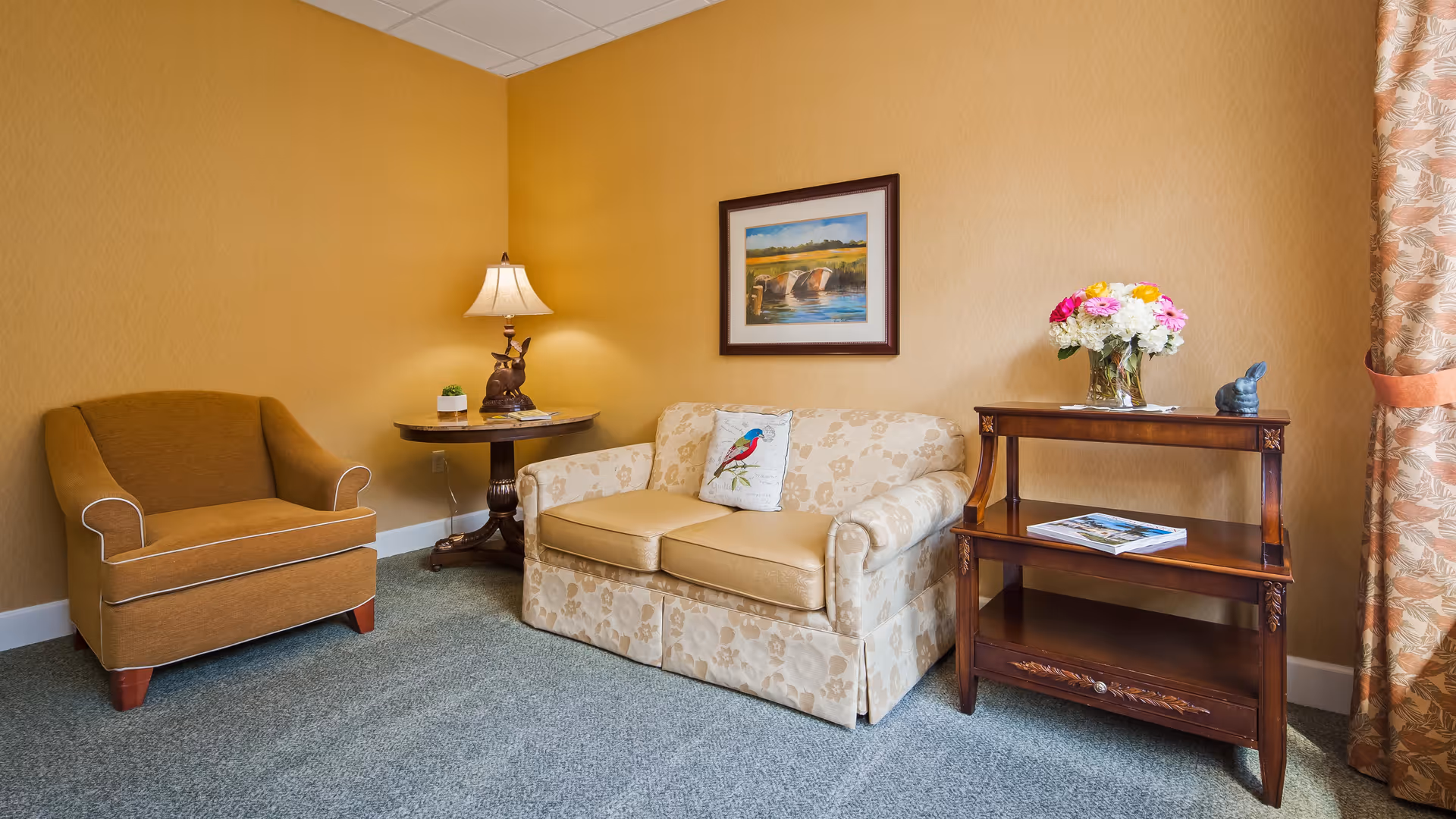 A cozy living room corner with a beige floral-patterned loveseat featuring a decorative bird pillow, a brown armchair, a round wooden side table with a lamp and small plant, and a wooden shelving unit holding a vase of colorful flowers and a book. The walls are painted a warm yellow, and a framed landscape painting hangs above the loveseat. A curtain with a leaf pattern is partially visible on the right.