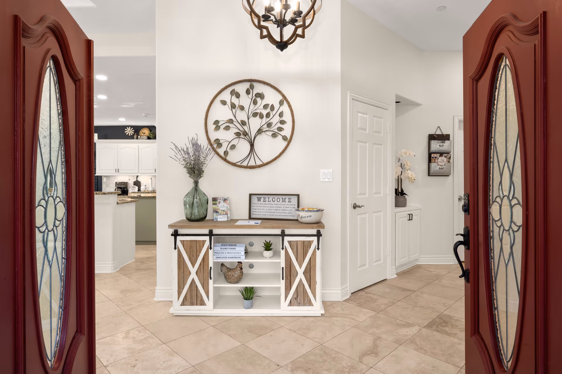 View through open double red doors with decorative glass panels into a bright interior space featuring a small wooden console table with sliding barn doors, decorated with a green vase with lavender, a welcome sign, and a bowl. Above the table is a round metal wall art piece shaped like a tree. The floor is tiled with beige tiles, and white walls and doors are visible, along with a glimpse of a kitchen area with white cabinets and granite countertops.