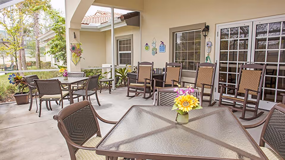 Outdoor patio area with several tables and chairs, including rocking chairs, decorated with a small vase of flowers on the table in the foreground. The patio is adjacent to a building with windows and a glass door, surrounded by greenery and trees.