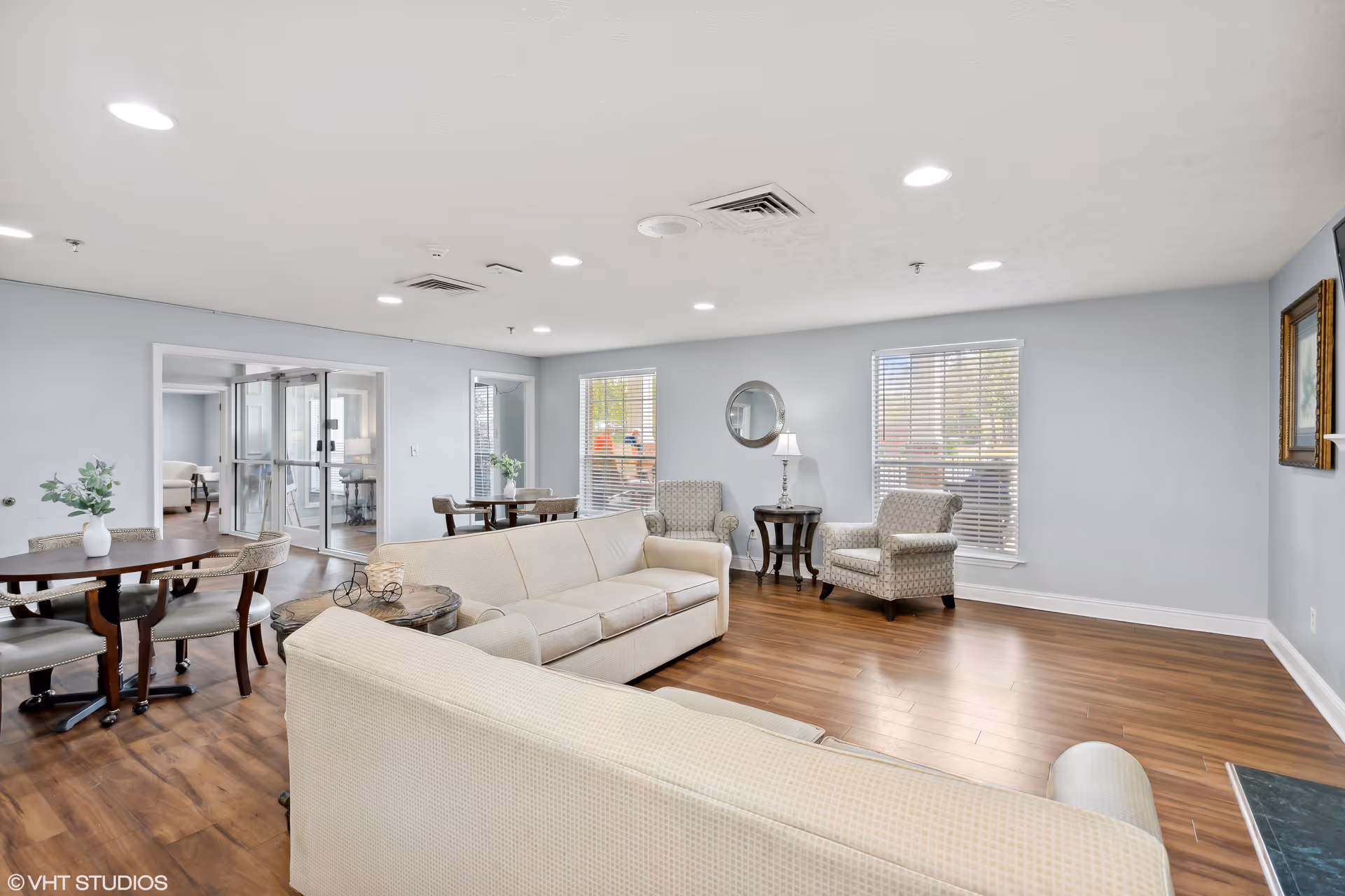 A bright and spacious living room area with light-colored sofas, two patterned armchairs, a round mirror on the wall, a side table with a lamp, and two windows with blinds. There are also two round tables with chairs, wooden flooring, and a doorway leading to another room.