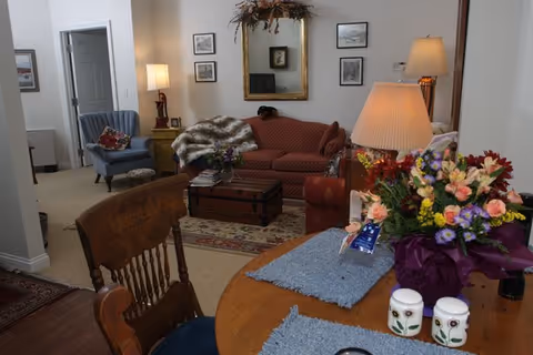 Interior living room and dining area with a wooden table holding flowers and placemats, upholstered sofa and armchairs, lamps, and framed pictures.