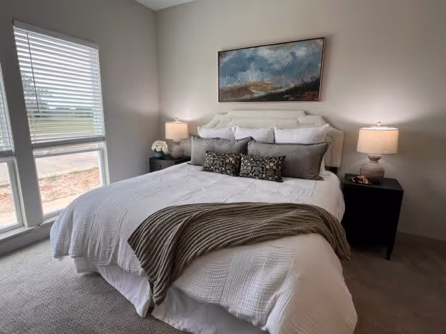 Light-filled bedroom with a neatly made bed in white linens, decorative pillows, bedside tables and lamps, wall art above the headboard, and a large window to the left.