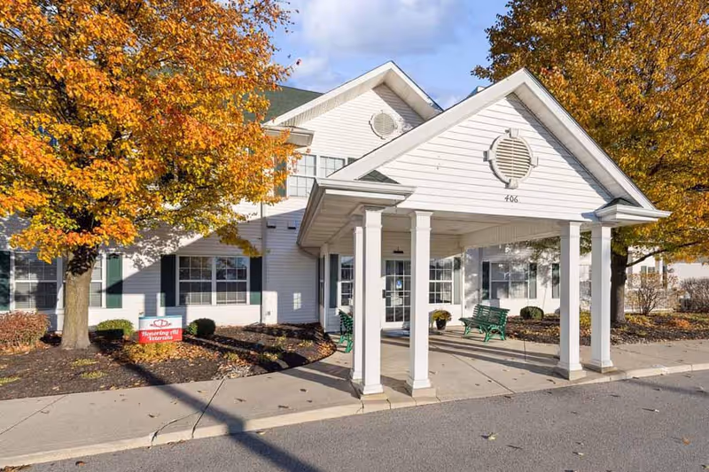 Exterior front view of a white senior living facility building with a covered entrance supported by four white columns. There are green benches under the entrance canopy and autumn-colored trees with orange and yellow leaves surrounding the building. A small sign near the tree reads 'Honoring all Veterans.'