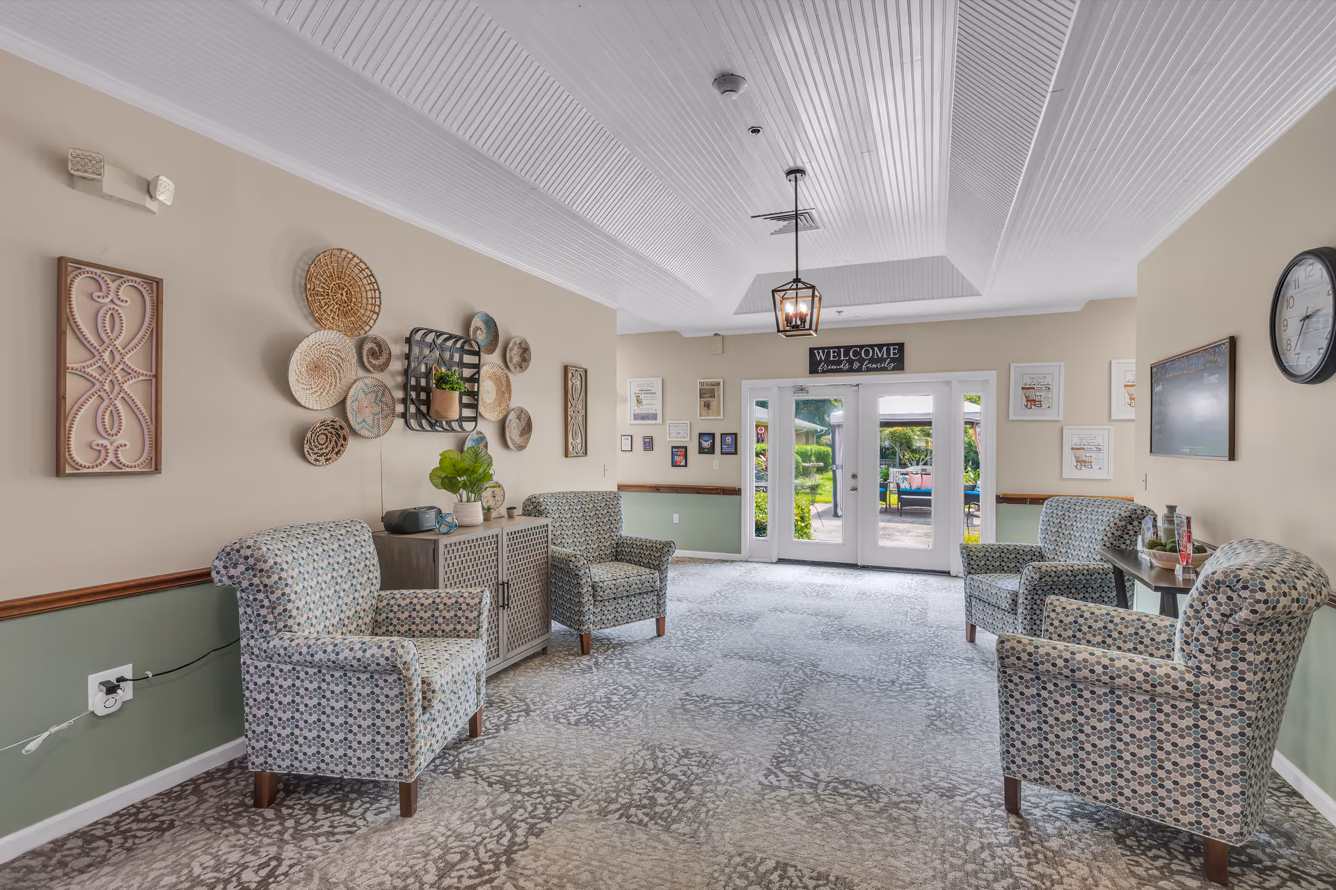 A bright and welcoming sitting area with four patterned armchairs arranged around a small table. The walls are decorated with woven baskets and framed artwork. Double glass doors at the far end open to a garden patio area. A clock and a welcome sign are visible on the walls.