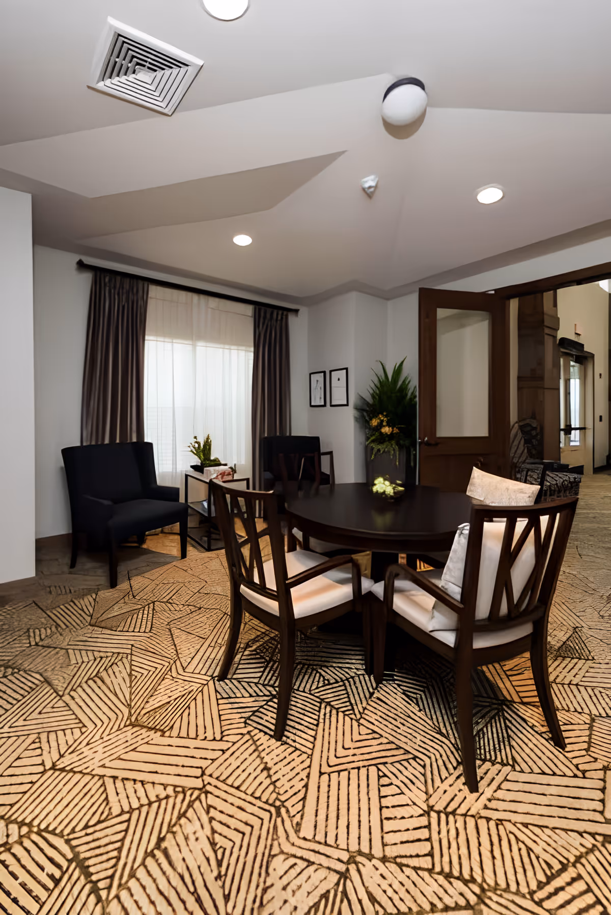 A cozy common room with a round dark wood table and chairs, armchairs by a curtained window, and a patterned carpeted floor.