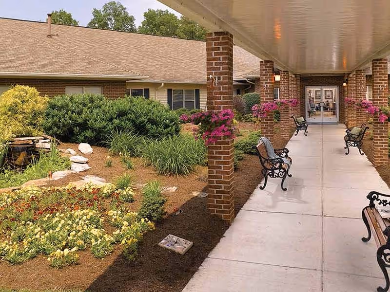 Covered walkway with benches and hanging flower baskets leading to the entrance of a single-story senior living building with landscaped gardens.
