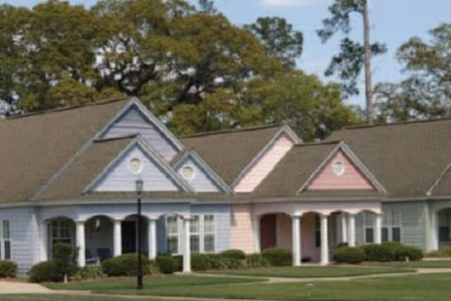 Exterior view of a single-story senior living facility building with pastel-colored sections in blue, pink, and light green, featuring gabled roofs, white columns, and well-maintained landscaping with bushes and a lawn.