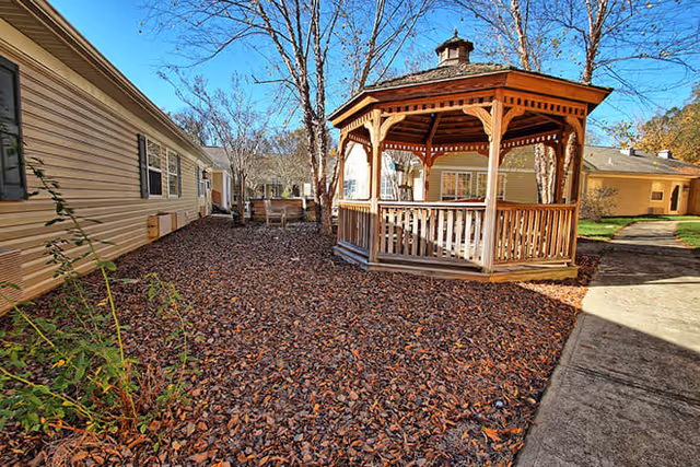 Outdoor area of a senior living facility featuring a wooden gazebo surrounded by buildings with beige siding and leaf-covered ground. There are leafless trees and a concrete pathway leading past the gazebo.