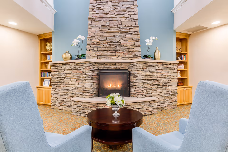 Cozy common room with a central stone fireplace flanked by built-in shelves, two blue armchairs, and a round table with flowers.