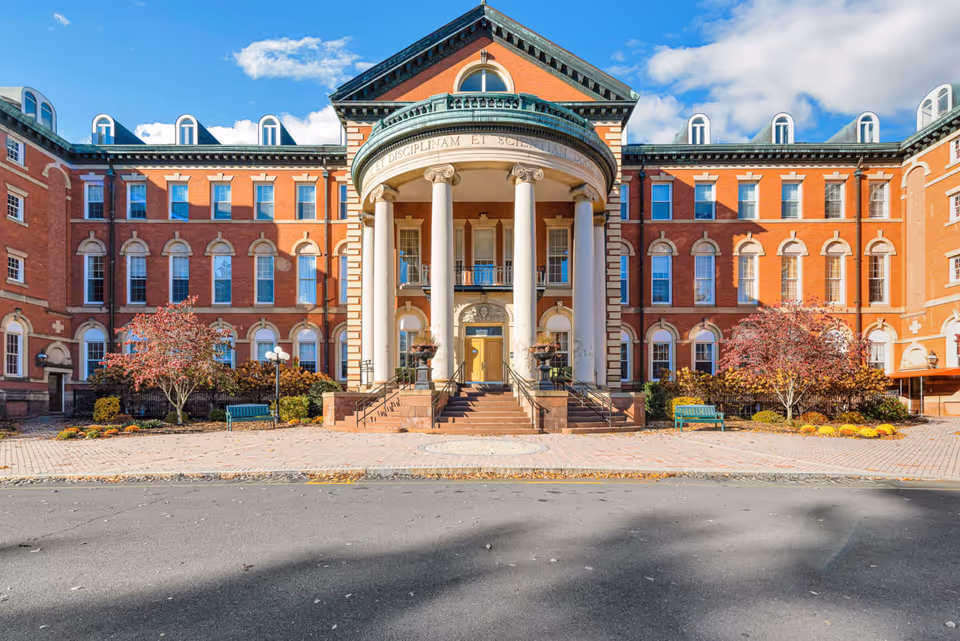 Front exterior view of a large, historic brick building with tall white columns supporting a rounded portico entrance. The building has multiple windows with decorative arches, a green roof trim, and a paved area with benches and small trees in front. The sky is blue with some clouds.