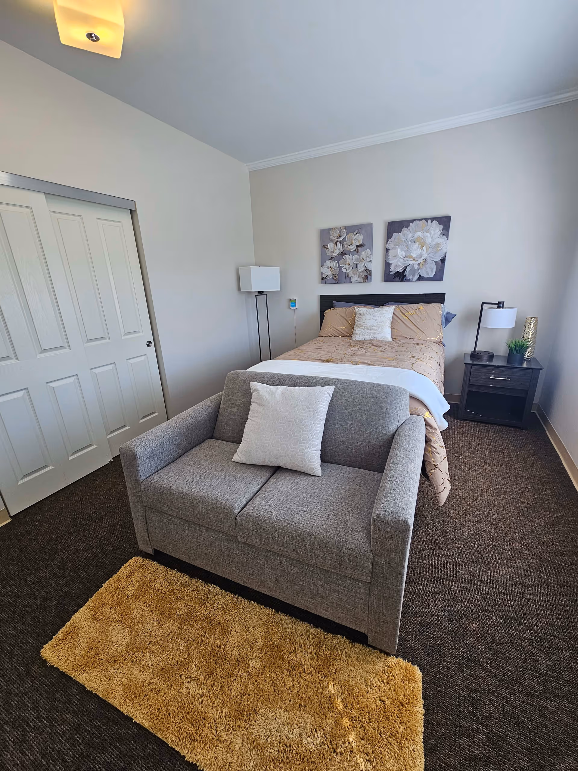 A bedroom with a bed covered in beige and white bedding, two floral paintings above the headboard, a gray loveseat with a white pillow in front of the bed, a beige rug on the dark carpeted floor, a nightstand with a lamp and small plant, a floor lamp, and a closed white sliding closet door.